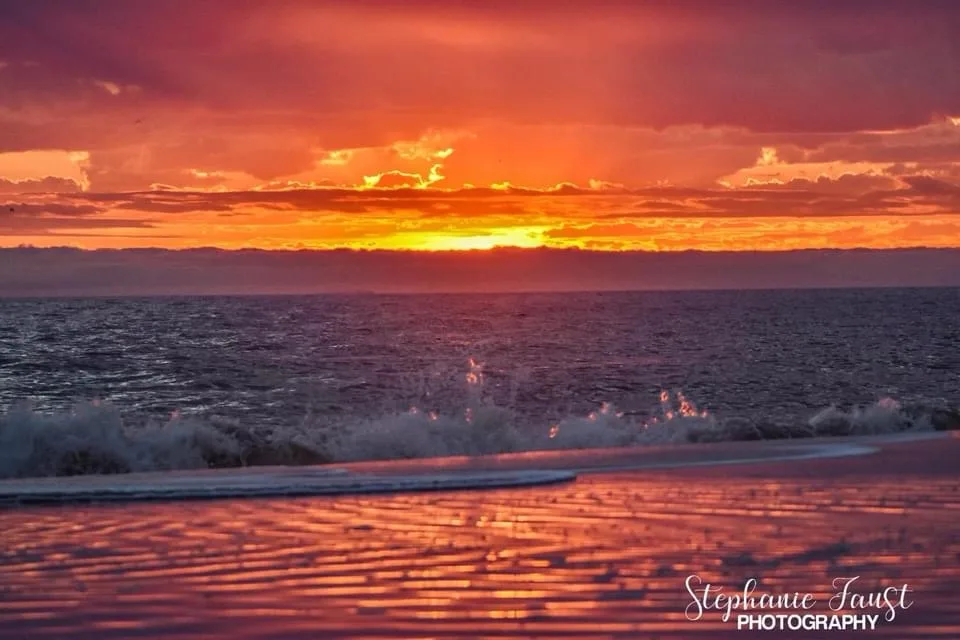 A vibrant sunset over the ocean with fiery orange and red clouds, waves crashing onto a wet sandy beach reflecting the sky's…