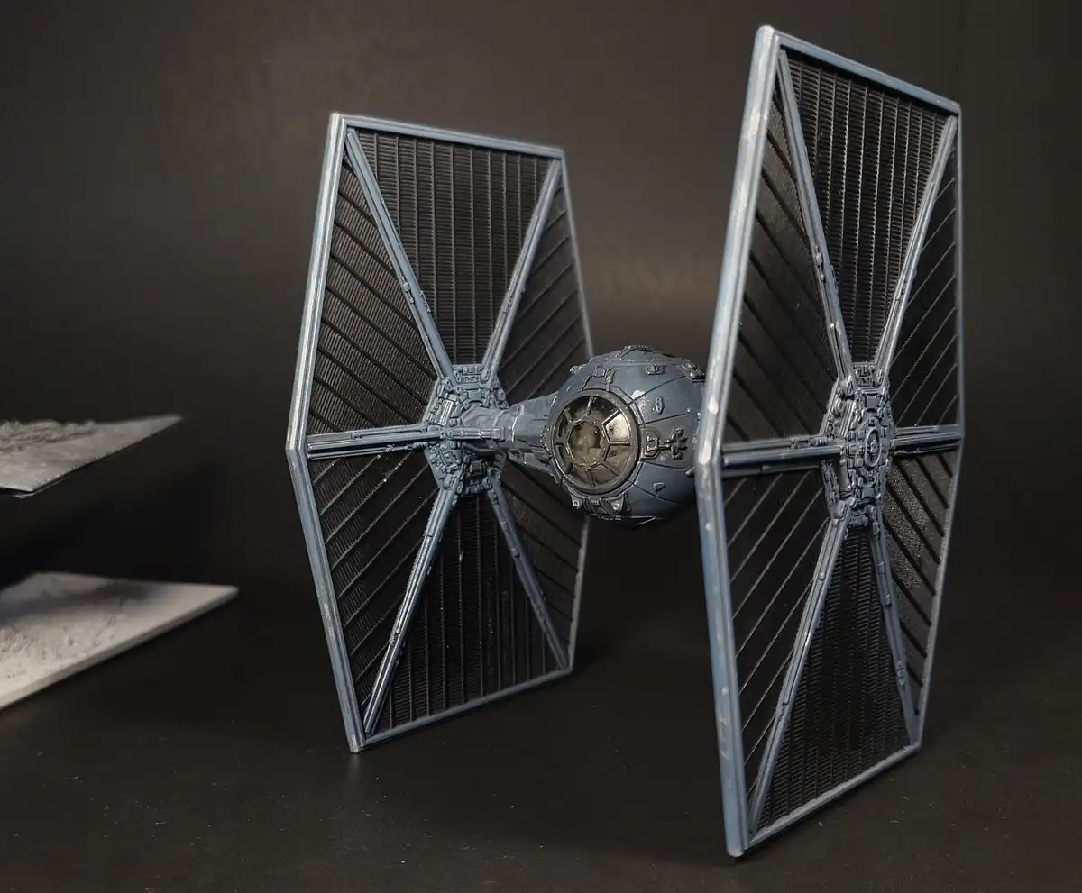 A gray, unpainted TIE fighter model stands upright on a dark surface, its wings angled outward. In the background, other…