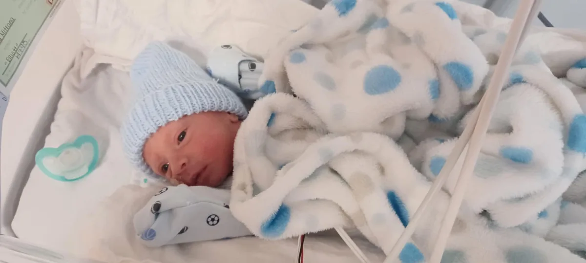 A newborn baby, wearing a light blue knit cap and wrapped in a white blanket with blue polka dots, lies in a hospital…