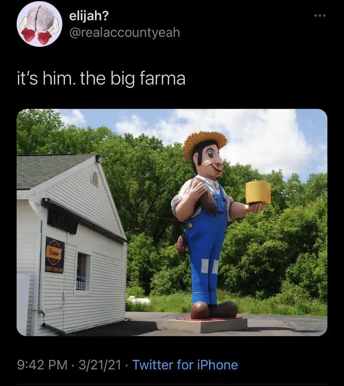 A large, cartoonish farmer statue wearing blue overalls and a straw hat stands outside a small white building labeled…