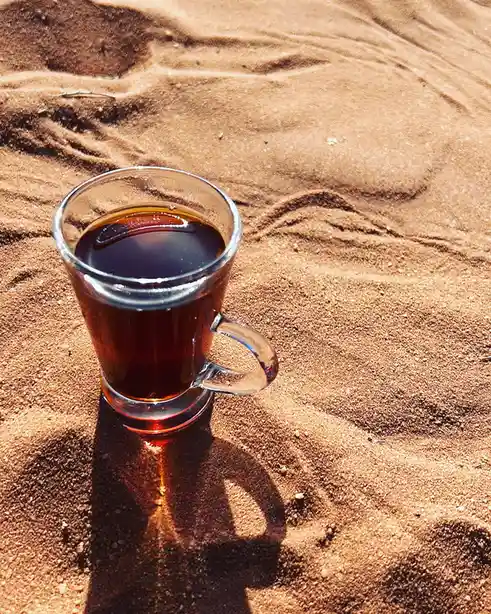 A clear glass mug filled with dark coffee sits on textured, sun-baked sand, casting a distinct shadow. The overlaid text…