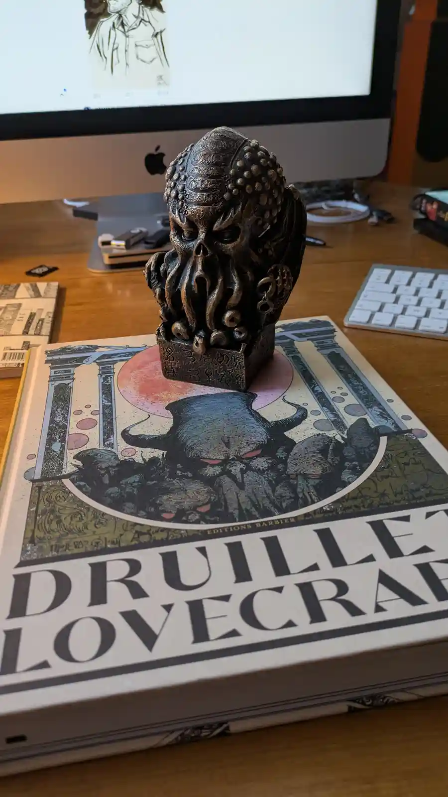 A dark bronze statue of Cthulhu sits atop a large art book titled 'Druillet Lovecraft' on a wooden desk, with a computer…
