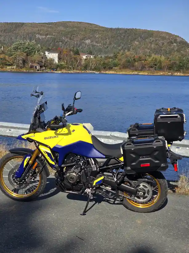 A bright yellow and blue Suzuki adventure motorcycle with black panniers is parked on a roadside next to a guardrail…