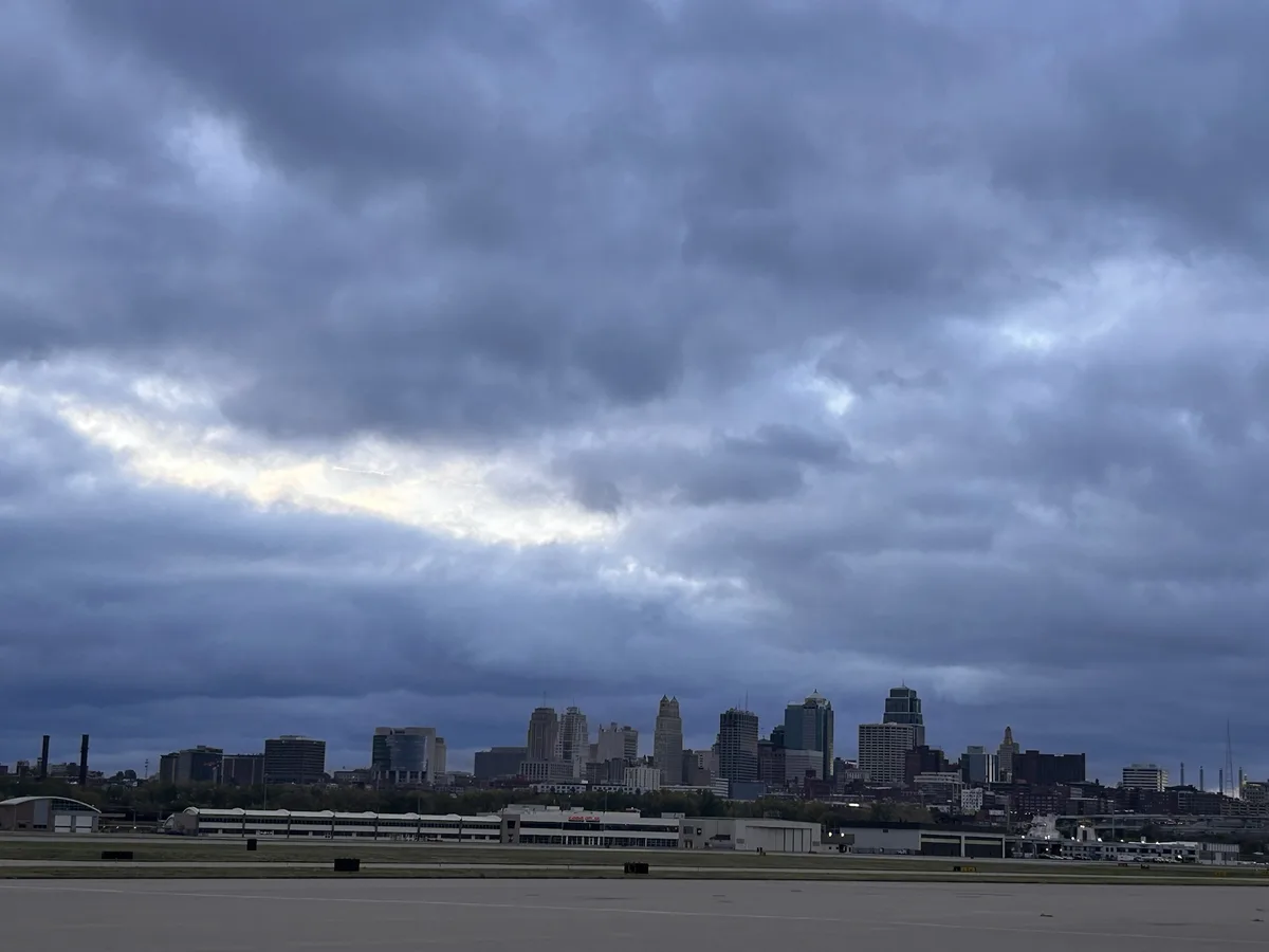 A photo of a city skyline under a heavy, overcast sky with a narrow band of sunlight breaking through the clouds. The…