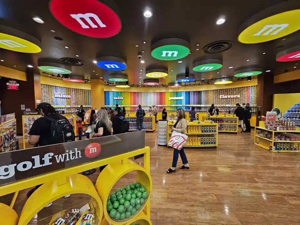The image shows the interior of an M&M's store with colorful circular 'm' logos on the ceiling, shelves stocked with candy…