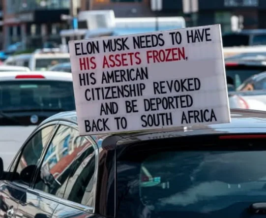 A protest sign is taped to the roof of a black car in a busy street, demanding that Elon Musk have his assets frozen, his…