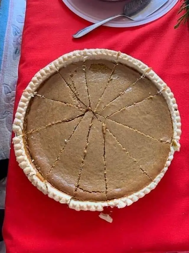 An overhead photo shows a whole pumpkin pie with 12 evenly cut slices on a red tablecloth, with a serving plate and spoon…