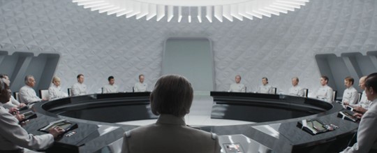 a wide shot from behind a person with short dark hair, looking at a large circular table surrounded by many people in white…