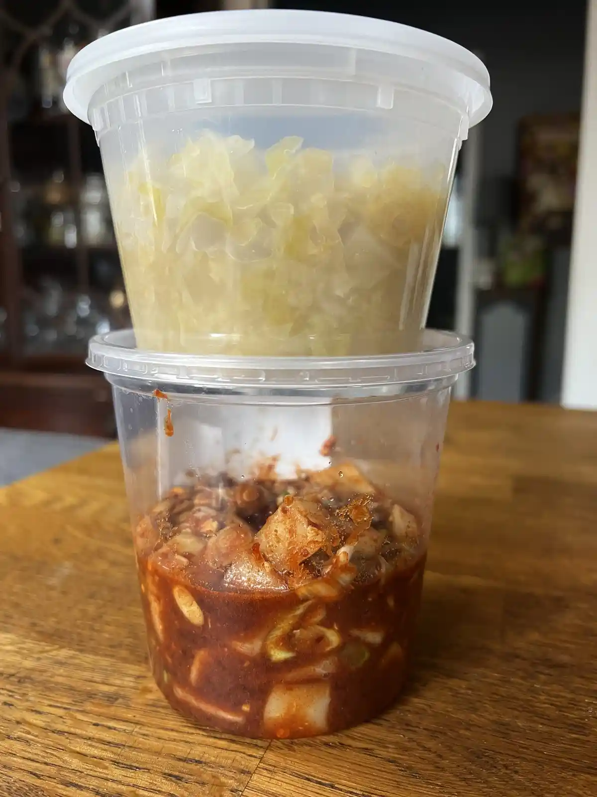 Two stacked clear plastic containers sit on a wooden table; the bottom one holds red, spicy-looking kimchi with visible…