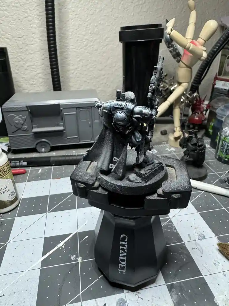 A heavily weathered, unpainted miniature of a space marine stands on a Citadel brand base, surrounded by hobby tools and…