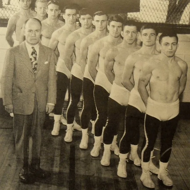 A vintage black-and-white photo shows a stern man in a suit standing beside a line of shirtless, muscular men in boxing…