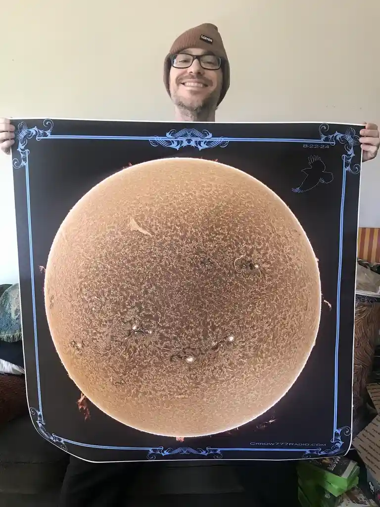 A smiling man wearing glasses and a brown beanie holds up a large poster depicting a detailed, textured image of the sun…