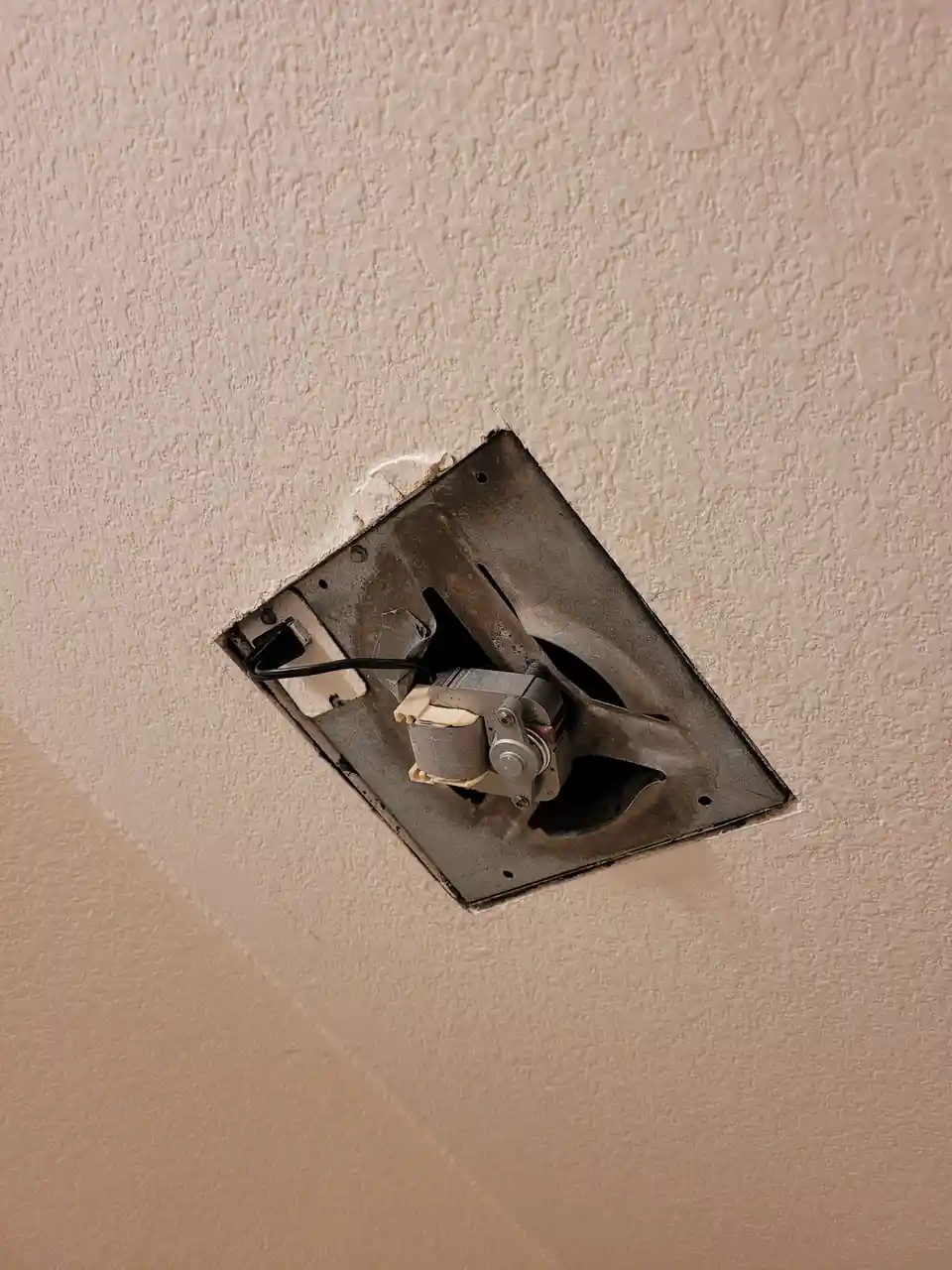 A close-up photo shows a square hole in a textured beige ceiling, revealing the exposed motor and blades of a ceiling fan…