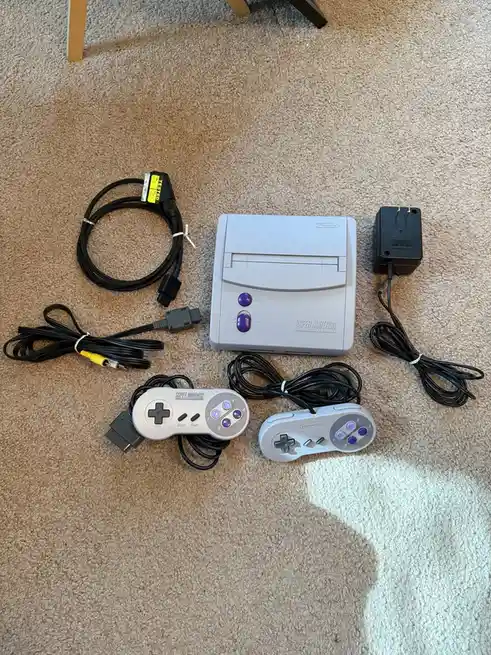 A complete Super Nintendo Entertainment System (SNES) console, two controllers, power adapter, and various cables are laid…