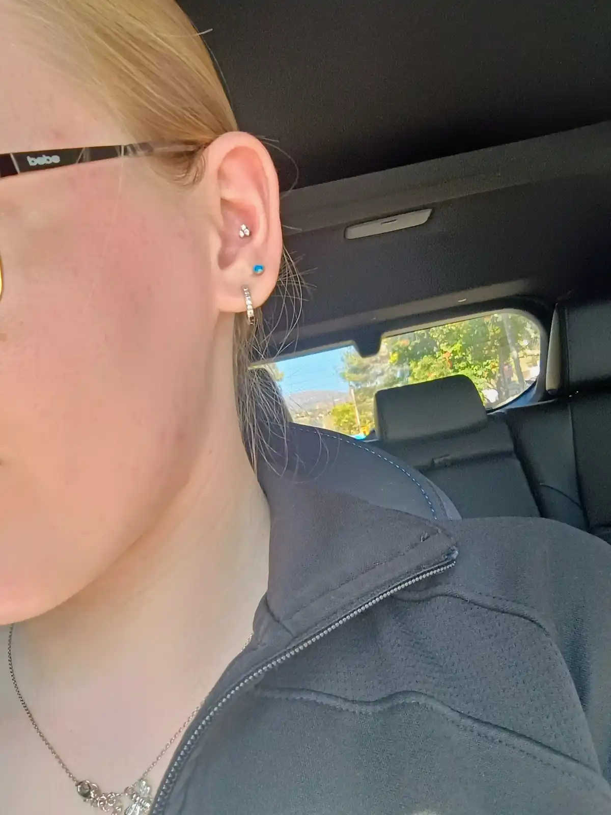 A close-up photo of a person's ear inside a car, showing multiple piercings including a stud, a blue gem, and a hoop. The…