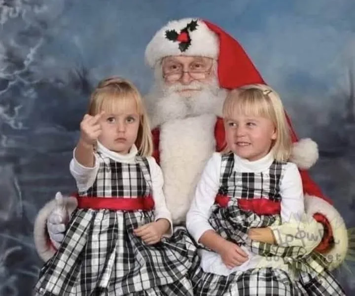 A posed Christmas photo shows Santa Claus seated between two young blonde girls in matching black-and-white plaid dresses…