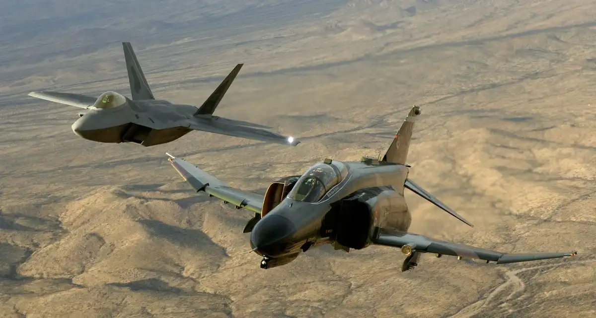 Two fighter jets fly side-by-side over a desert landscape: a modern stealthy F-35 on the left and a classic F-16 on the…