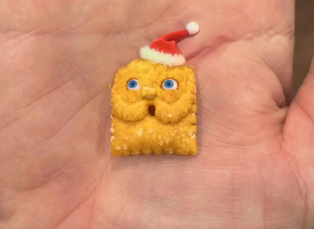 A small, square, yellow cracker with googly blue eyes, a tiny nose, and a mouth is held in a person's palm. It wears a…