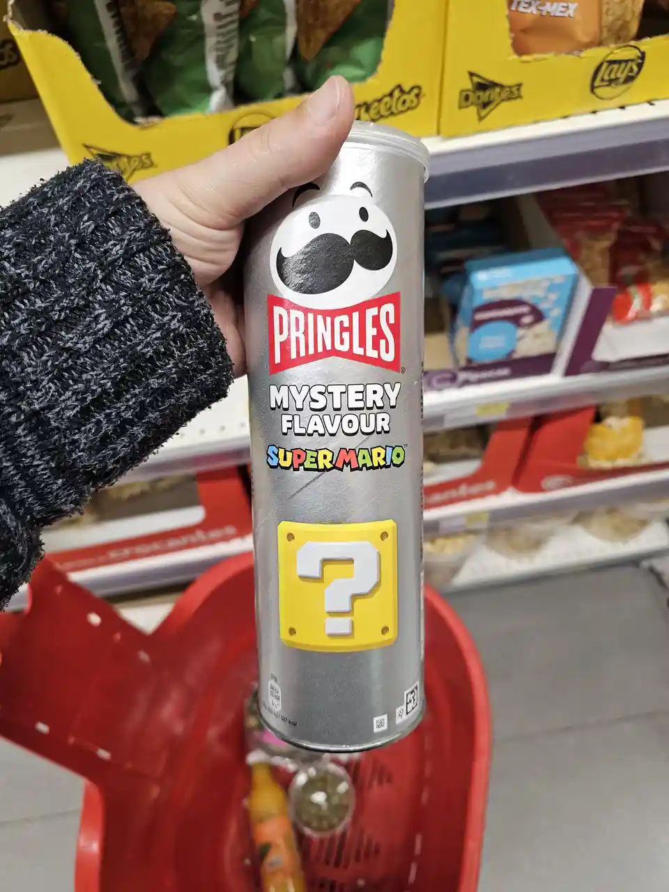 A hand holds a silver Pringles can labeled 'Mystery Flavour Super Mario' featuring a mustachioed face and a yellow question…