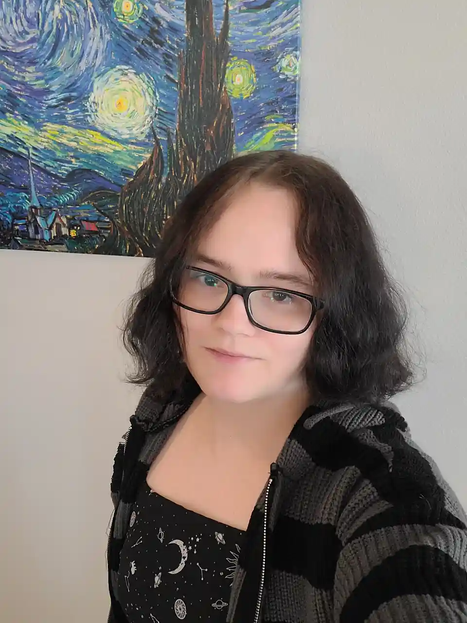 A person with dark hair and glasses stands in front of a wall, with a print of Vincent van Gogh's 'The Starry Night' visible…