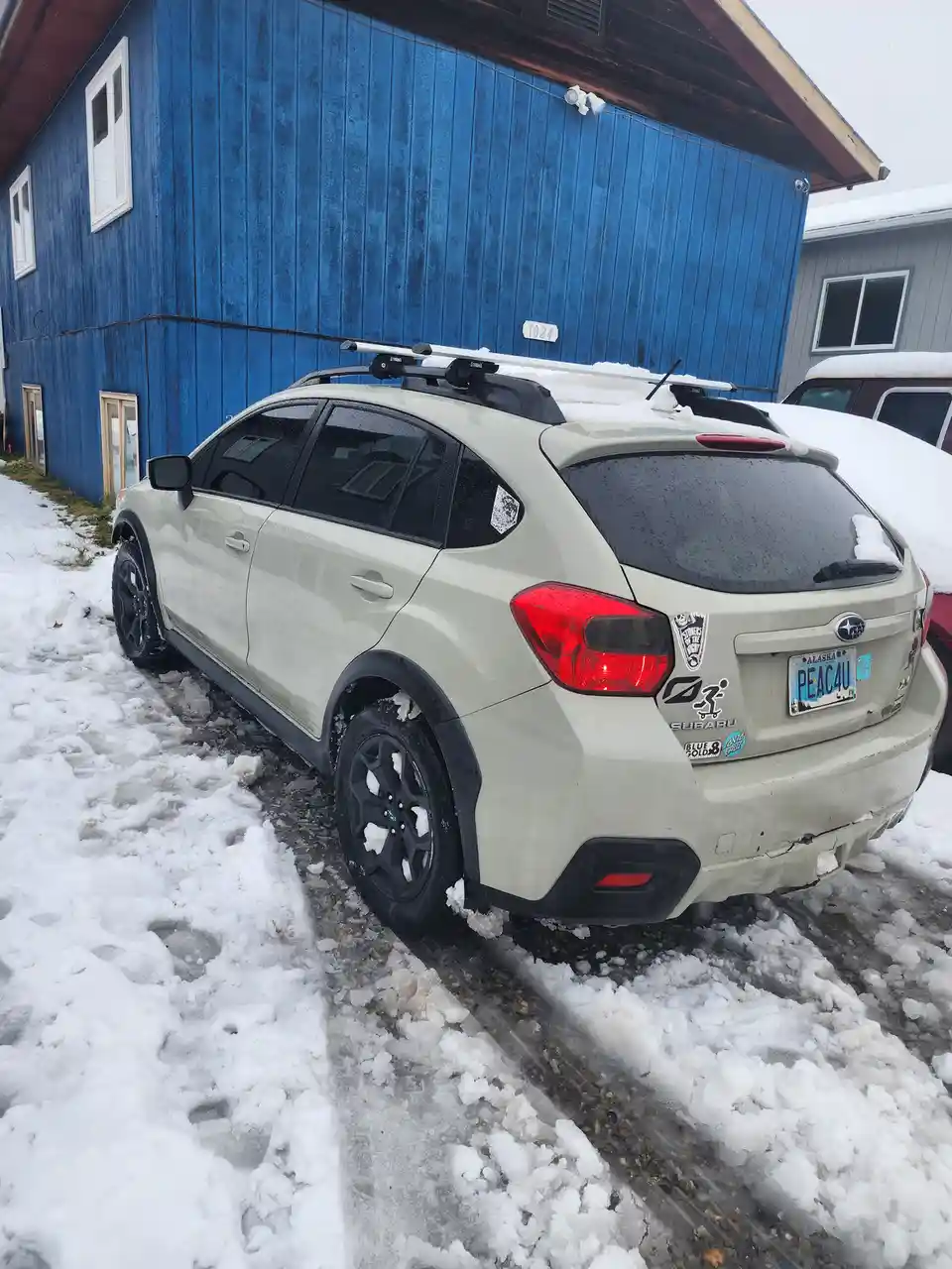 A light-colored Subaru SUV is parked in a snowy, icy driveway next to a blue wooden building. The car has a roof rack, black…