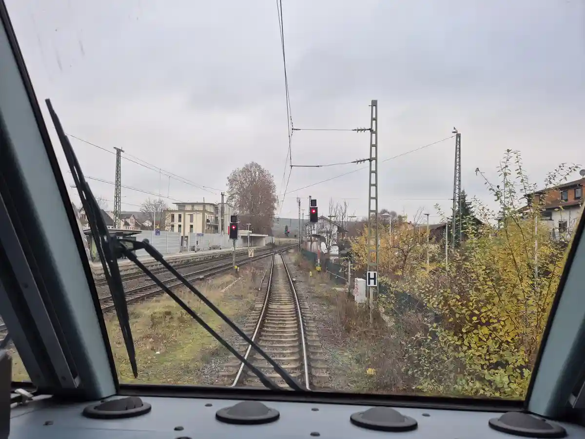A view from inside a train looking out at railway tracks approaching a station under an overcast sky, with a red traffic…