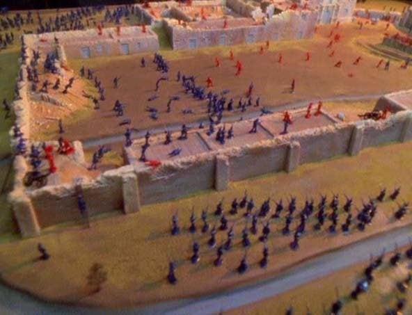 A miniature wargaming scene depicts a battle with blue and red plastic soldier figures on a diorama battlefield with stone…