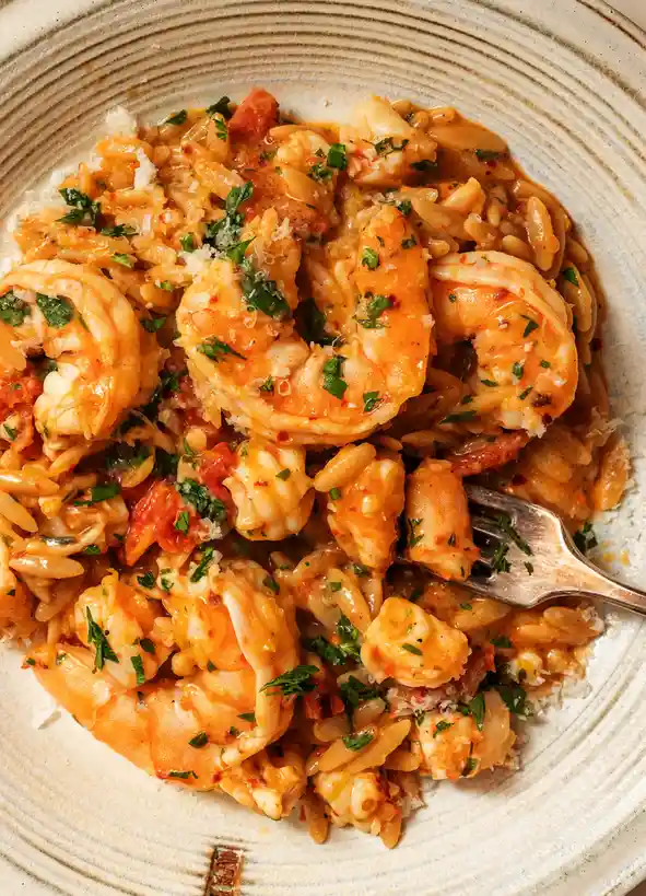 A close-up photo of a plate of orzo pasta mixed with large cooked shrimp, garnished with chopped parsley and grated cheese…