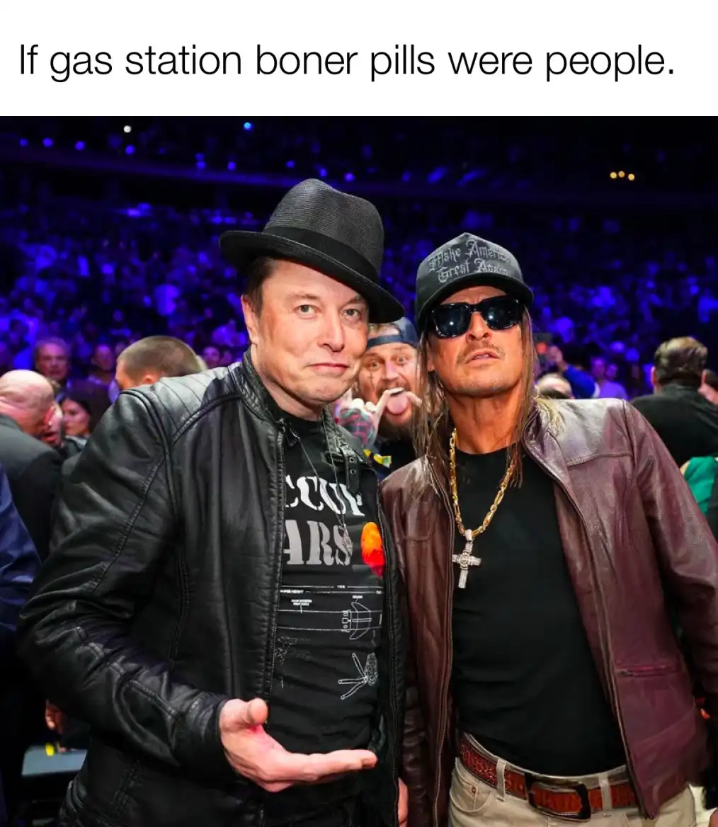 Two men stand side by side in a crowded venue with blue lighting; the man on the left wears a black fedora and leather…