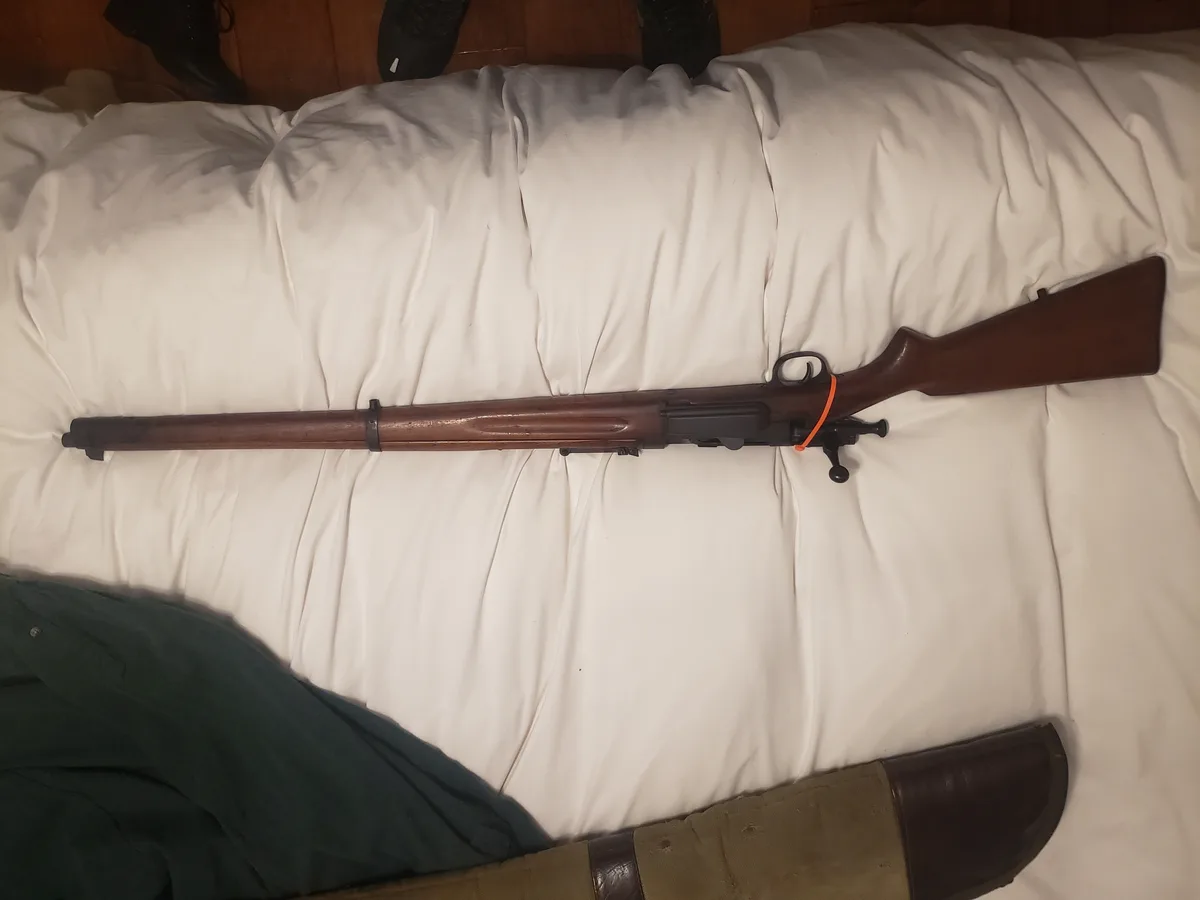 A wooden-stocked rifle with an orange safety strap lies horizontally across a rumpled white bedsheet. At the top of the…