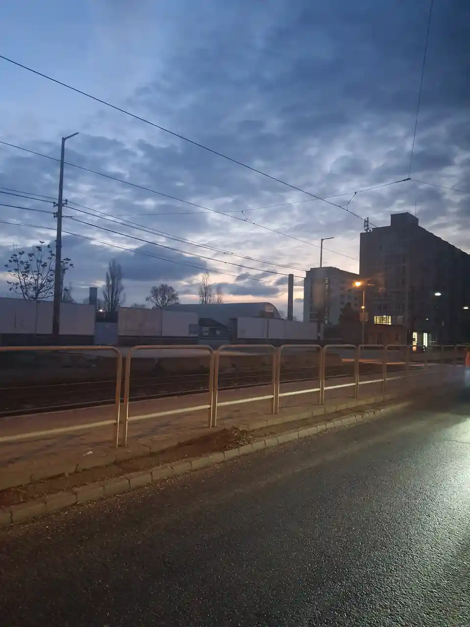 A photo of a dimly lit urban street at dusk with tram tracks, a metal railing, and apartment buildings under a cloudy sky…
