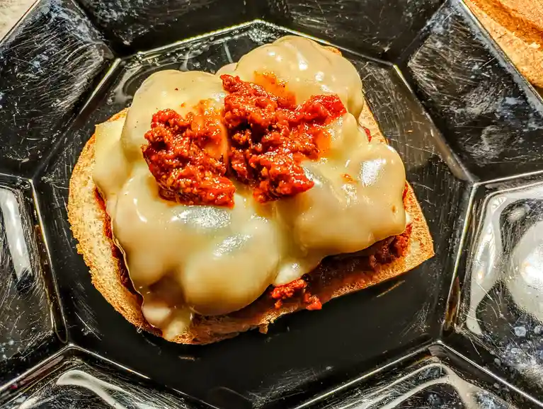 A close-up photo shows a single slice of bread topped with melted cheese and a dollop of red sauce, served on a black…