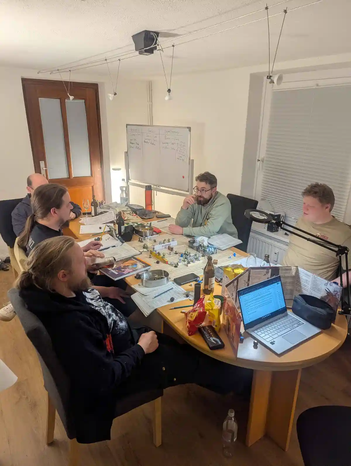 Four men are gathered around a cluttered wooden table engaged in a tabletop role-playing game, surrounded by miniature…