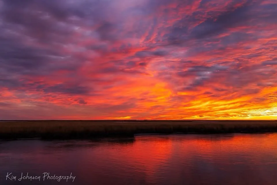 A breathtaking photograph of a vibrant sunset over a body of water, with fiery orange, red, and purple clouds reflected in…
