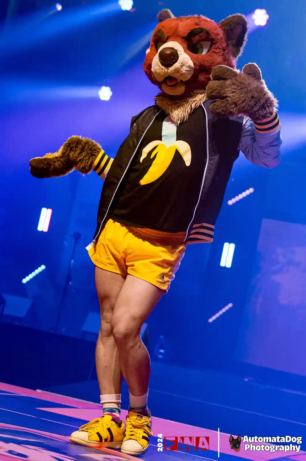 A person in a red and brown furry bear mascot costume strikes a pose on a stage under blue lights, wearing a black jacket…