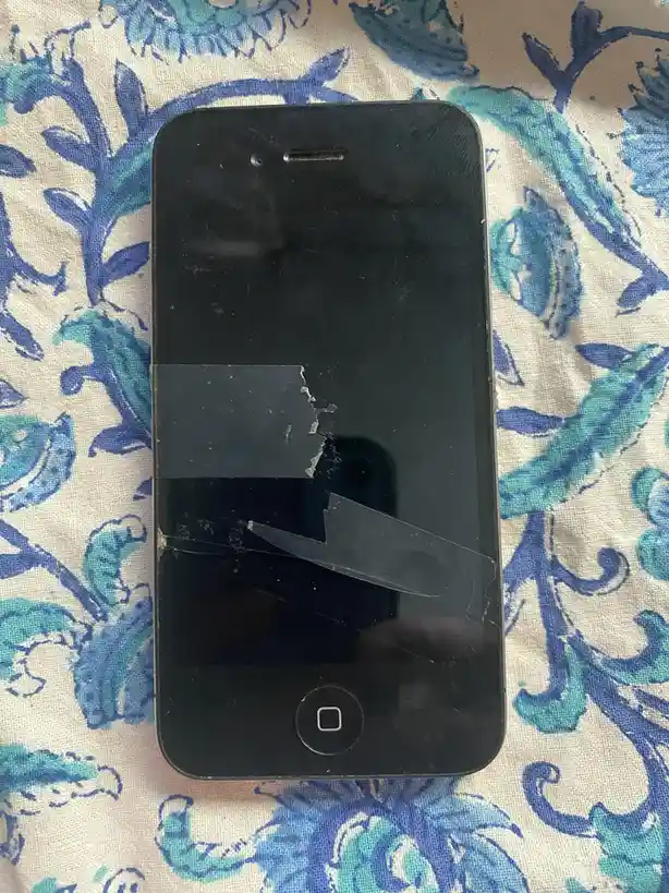 A black iPhone with a severely cracked and taped-up screen lies on a blue and white floral fabric. The overlaid text reads…