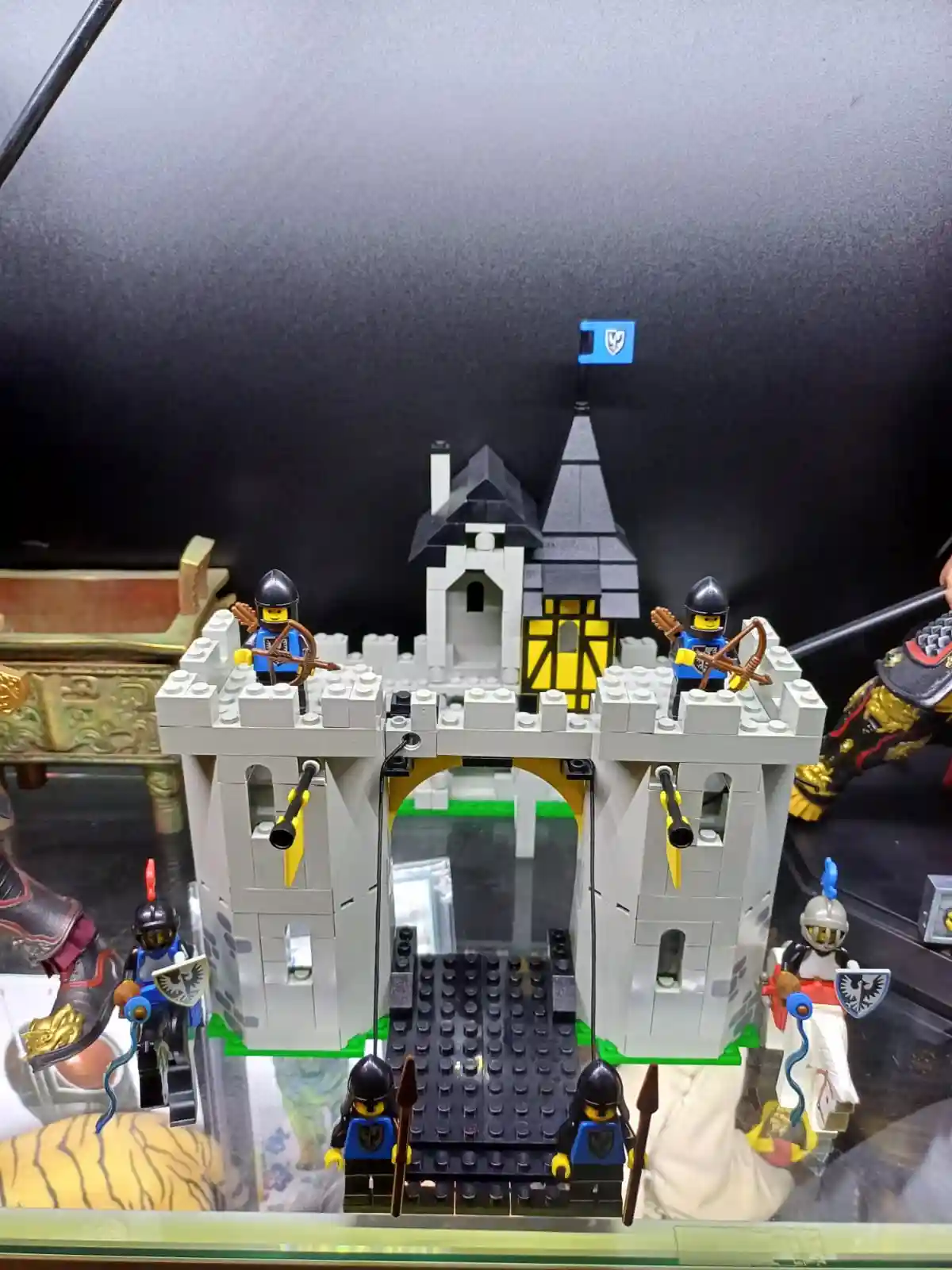 A Lego castle with gray walls, a black roof, and a blue flag, surrounded by Lego knights holding bows, spears, and shields…