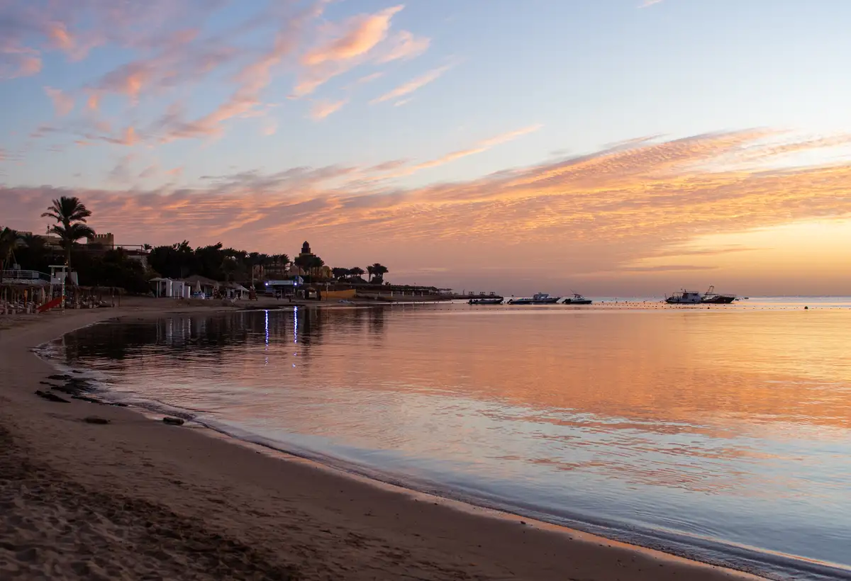 A serene beach at sunrise or sunset with calm water reflecting orange and pink clouds, boats anchored offshore, and…