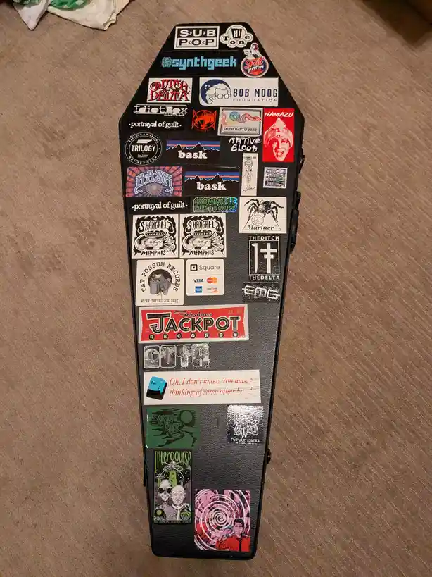 A black guitar case shaped like a coffin is covered in numerous stickers from music labels, festivals, and bands like Sub…