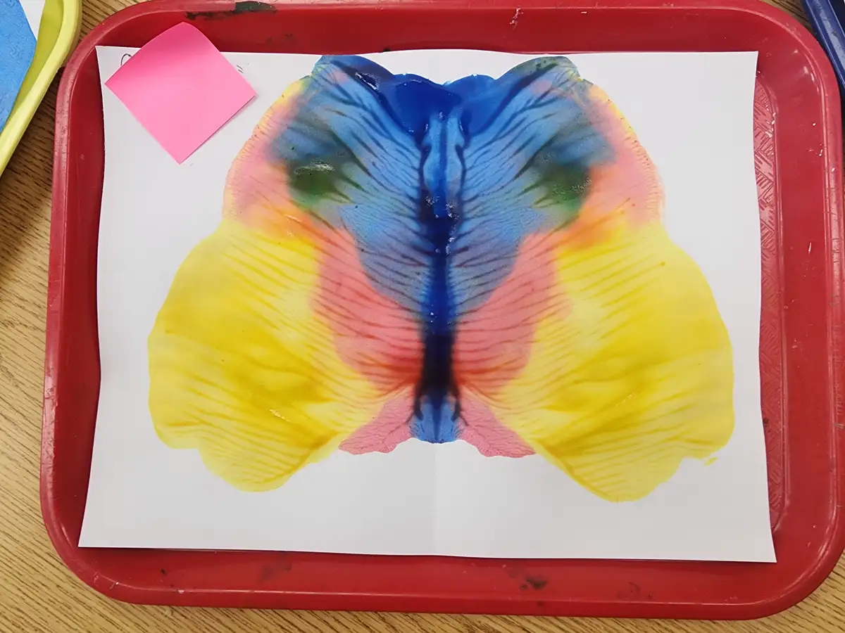 A child's art project shows a colorful, symmetrical butterfly made from folded paper and paint, resting on a red plastic…