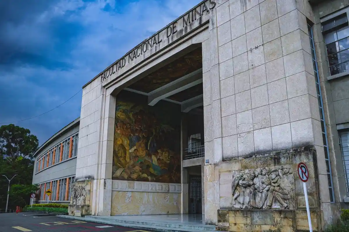 A wide-angle photo shows the grand stone facade of a university building labeled 'FACULTAD NACIONAL DE MINAS' with a large…