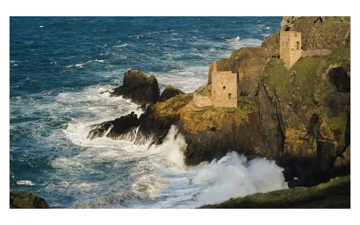 A scenic photo shows two stone towers of a ruined coastal fortress perched on a steep, grassy cliff overlooking a turbulent…