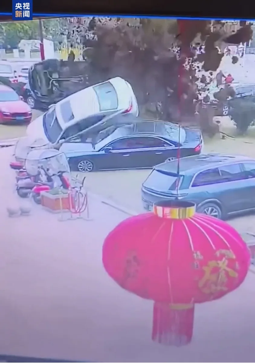 监控画面显示一辆白色轿车在停车场发生严重事故，车身翻转并撞上其他车辆，现场扬起大量烟尘。前景中有一个巨大的红色中式灯笼，与背景的混乱场面形成强烈对比。画面左上角有“央视新闻”的水印。