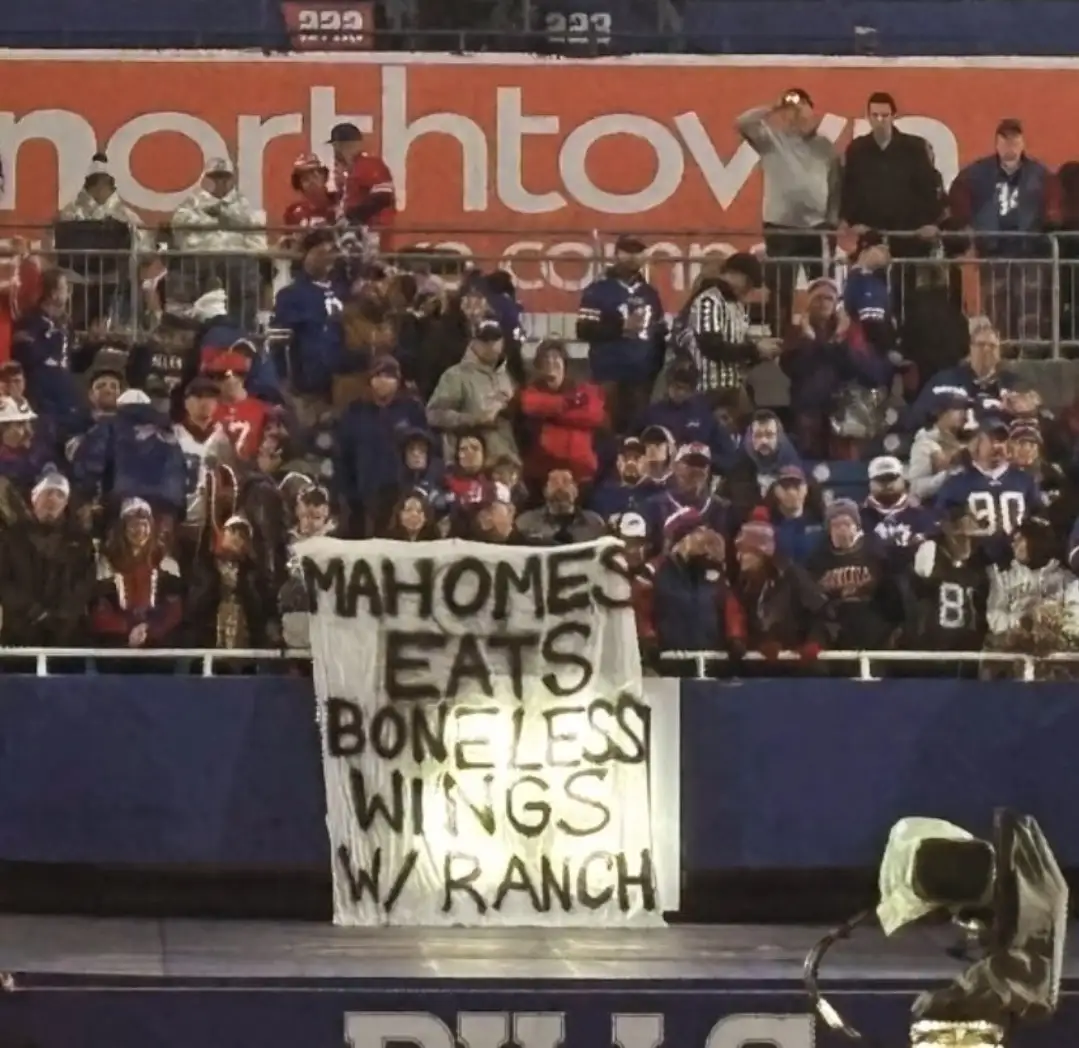 A crowd of football fans at a stadium holds up a handmade white banner with black marker text that reads 'MAHOMES EATS…