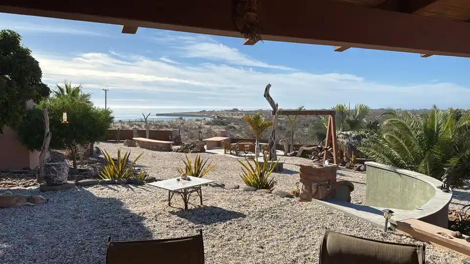 A serene outdoor patio with gravel ground, desert plants, a curved pool, and ocean view under a wooden overhang. A dog…