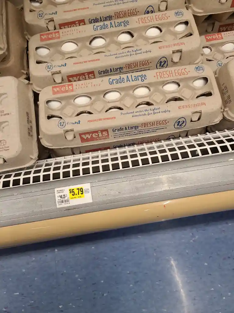 A close-up photo of egg cartons stacked on a refrigerated grocery store shelf, with a price tag showing $5.79 for a dozen…