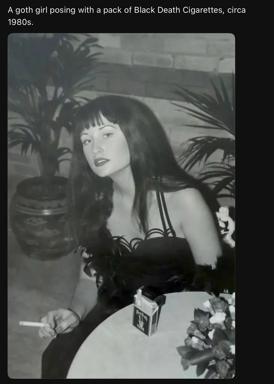A black-and-white photo from the 1980s shows a woman with dark hair and bangs, wearing a black top, sitting at a table. She…