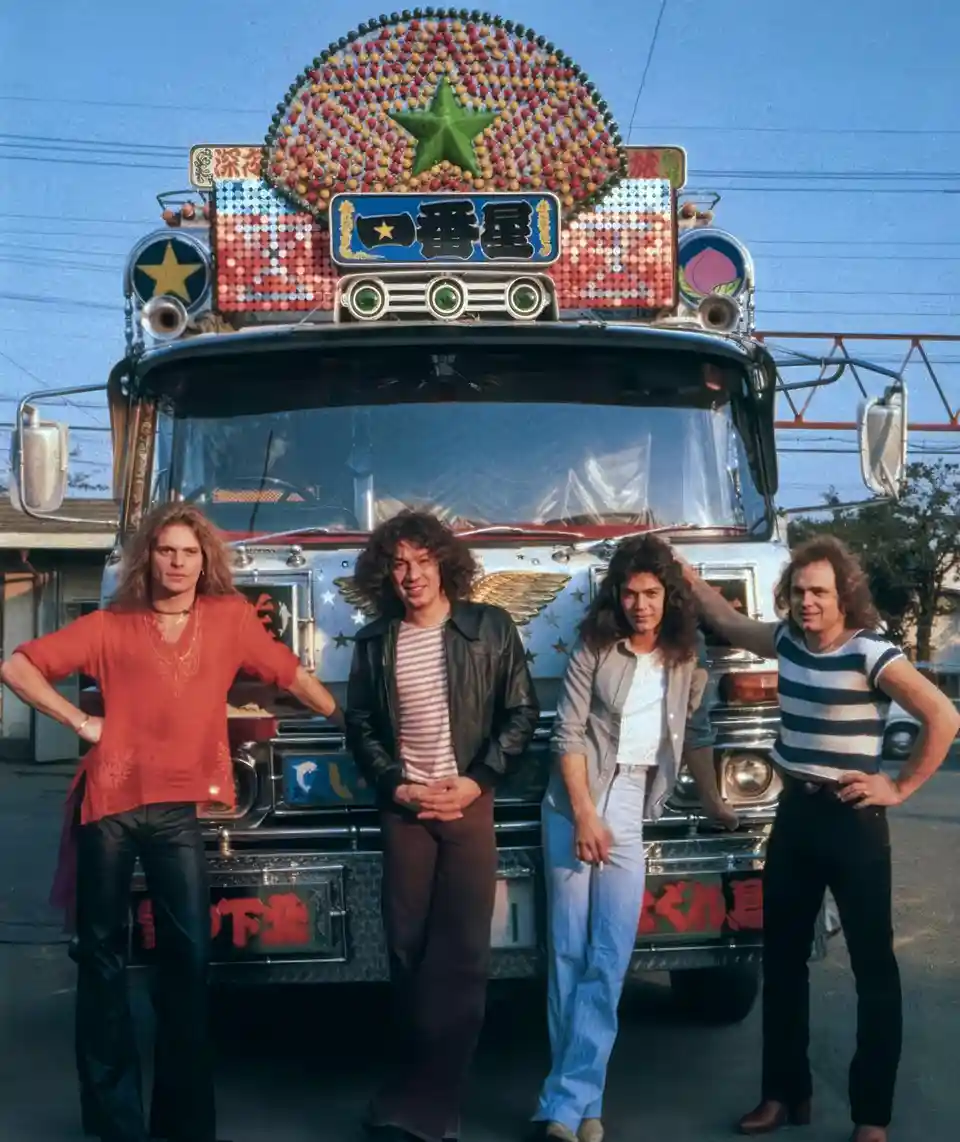 Four men with long hair pose confidently in front of a heavily decorated, ornate Japanese truck adorned with colorful beads…