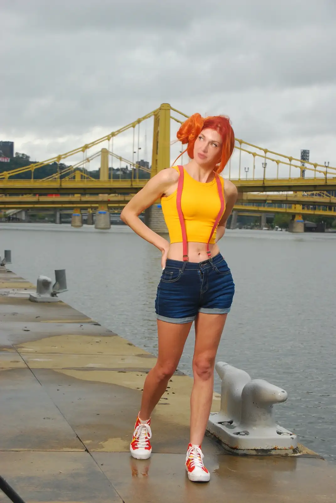 A woman with bright orange hair, wearing a yellow crop top, pink suspenders, denim shorts, and red-and-white sneakers, poses…