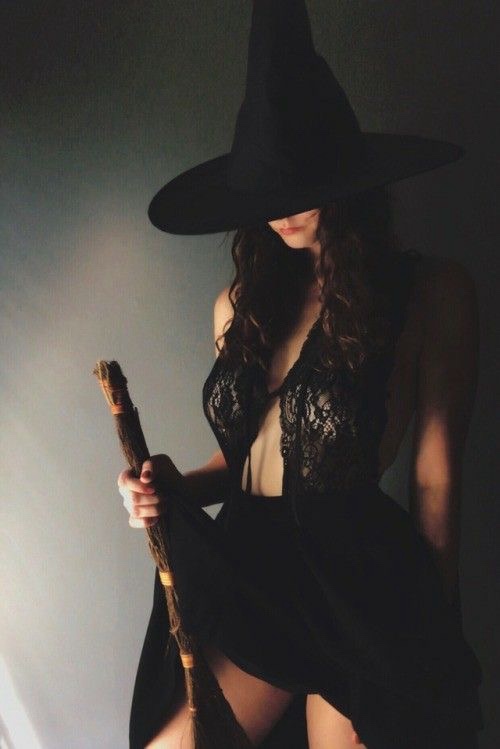 A woman in a black witch's hat and black lace outfit holds a broomstick, her face partially obscured by the hat. The…